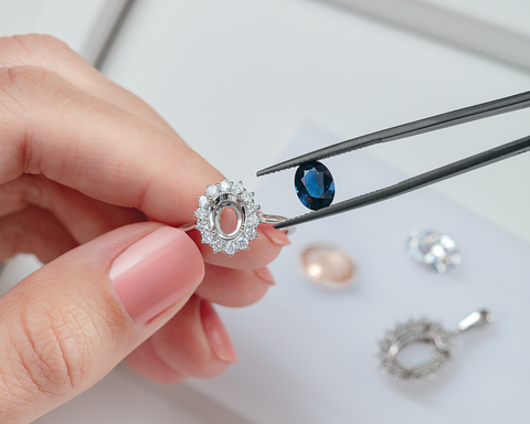 A close-up of a hand holding a silver halo ring with a blue oval gemstone, while black tweezers hold a matching blue stone. Other loose gemstones and a silver halo setting are visible on a white tray in the background.